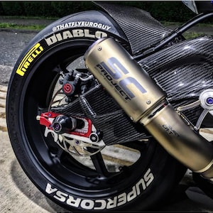 May include: A motorcycle's rear wheel, featuring a Pirelli Diablo tyre with white lettering. A gold exhaust pipe with "SC Project" text is visible. The bike has carbon fibre components. The wheel is black.