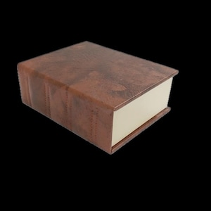 Op de afbeelding: Een bruine, op een boek lijkende doos van imitatieleer met een crèmekleurig interieur. De doos heeft een textuuroppervlak en gestikte details langs de rug, ontworpen om op een klassiek boek te lijken. De doos is gesloten en staat tegen een zwarte achtergrond.