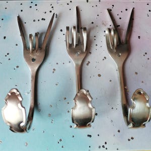 May include: Three silver-coloured forks repurposed as decorative wall hooks. Each fork is bent to form a hand gesture: "I love you", "middle finger", and "peace". The hooks have a decorative, curved base.