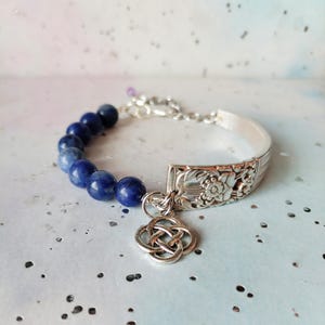 May include: A bracelet featuring a row of round, deep blue beads connected to a silver-toned metal segment with an embossed floral pattern. A silver-toned metal charm in the shape of an intricate, interlaced knot hangs from the junction.