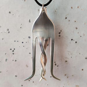 May include: A silver-coloured metal pendant shaped like a modified fork with two outer tines curving outwards and two inner tines twisted together in the centre. The pendant is suspended from a thin black cord, creating a unique jewellery piece.