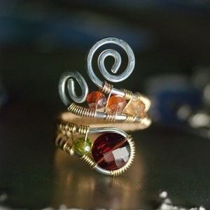 May include: A silver and gold wire wrapped ring with a large red gemstone and smaller orange and green gemstones.