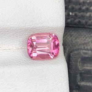 Puede incluir: Una gema rosa, talla cojín, de forma rectangular con esquinas redondeadas. Las facetas de la gema reflejan la luz. Se encuentra sobre una superficie blanca, con un fondo gris oscuro visible en el lado derecho.