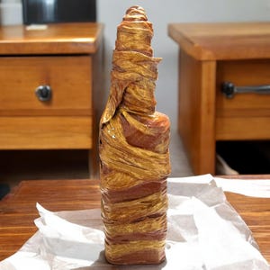 Könnte beinhalten: Eine Glasflasche, die in goldfarbenes und braunes Stoff gewickelt ist und einer Mumie ähnelt. Die Flasche steht auf einer weißen Oberfläche.