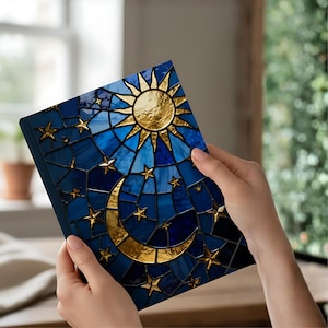 May include: A blue journal with a stained-glass design featuring a gold sun, moon, and stars. The cover has a dark blue border and a mosaic pattern in shades of blue. The sun and moon have a metallic gold finish.
