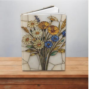 Puede incluir: Un diario abierto con un diseño floral. La portada presenta un ramo de margaritas, flores amarillas y azules y espigas de trigo. Las flores están sobre un fondo blanco con un patrón agrietado, similar a un mosaico. El diario descansa sobre una superficie de madera.