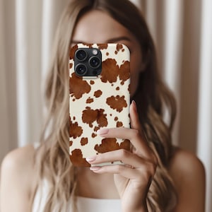 May include: A phone case with a cowhide pattern. The case features a cream-colored background with irregular brown spots, mimicking the look of cowhide. The case has a camera cutout and is held in front of a person.