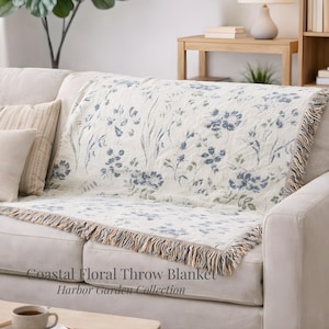 Puede incluir: Una manta crema con un estampado floral azul y flecos está sobre un sofá. La manta tiene el texto "Coastal Floral Throw Blanket" y "Harbor Garden Collection".