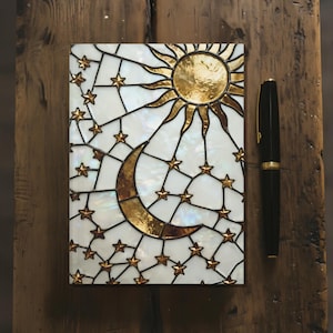May include: A decorative journal with a celestial design featuring a gold sun, crescent moon, and stars on a white background. A black pen with gold accents rests beside the journal on a wooden surface. The journal's cover has a stained glass appearance.