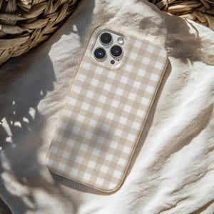 Peut inclure: Coque de téléphone à motif vichy beige et blanc. L'étui a une bordure transparente autour des lentilles de la caméra et repose sur un tissu beige. L'étui du téléphone est de forme rectangulaire avec des coins arrondis.