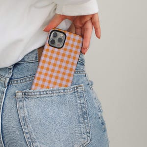 Peut inclure: Coque de smartphone à motif vichy orange et lavande. L'étui est partiellement visible dans la poche arrière d'un jean en denim délavé. Le boîtier du téléphone a une bordure noire.