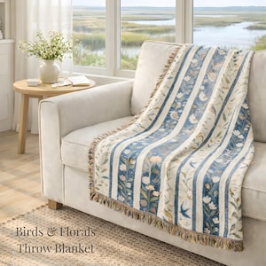 May include: A throw blanket with a striped pattern of blue and cream vertical stripes, adorned with floral and bird motifs. The blanket has a fringed edge and is draped over a cream-colored sofa. The text "Birds & Florals Throw Blanket" is visible.