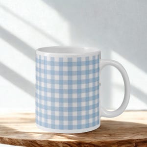 Może przedstawiać: Biały ceramiczny kubek z jasnoniebieskim i białym wzorem w kratkę vichy. Kubek ma zakrzywiony uchwyt i jest ustawiony na drewnianej powierzchni. Tło jest jasne z cieniami.