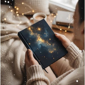 Op de afbeelding: Een donkerblauw notitieboek met een hemels ontwerp met gouden sterren en wolken. Het notitieboek is opengeklapt en toont de gedetailleerde illustraties op de omslag. Een zwart lint als bladwijzer is zichtbaar. Een perfect cadeau voor schrijvers en dromers.