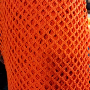 May include: Orange mesh fabric with a diamond pattern. The fabric is rolled up and the texture of the mesh is visible.
