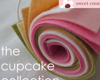 Puede incluir: Un rollo de tela de fieltro en varios colores pastel, incluyendo rosa, verde, amarillo y blanco. El fieltro está enrollado y atado con una cuerda blanca. El texto "the cupcake collection" está escrito en blanco debajo del rollo de fieltro. El texto "sweet emma jean" está escrito en blanco sobre una flor rosa con un centro rojo.