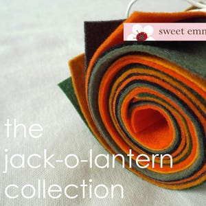 May include: A roll of felt fabric in various shades of orange, green, and brown. The felt is rolled up and tied with a white ribbon. The text "sweet emma jean" is printed on a white tag attached to the roll. The text "the jack-o-lantern collection" is printed below the roll of felt.