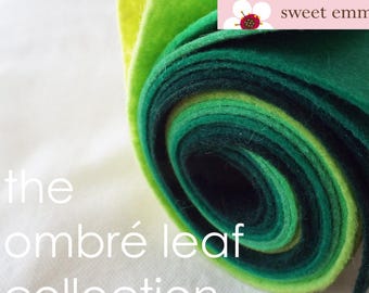 May include: A roll of felt fabric in shades of green, from light to dark, with the text "sweet emma jean" on a pink label. The felt is rolled up and the ends are visible. The text "the ombré leaf collection" is written below the roll of felt.