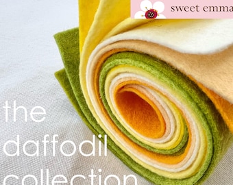 Puede incluir: Un rollo de tela de fieltro en tonos de amarillo, verde y blanco. El fieltro está enrollado y los extremos son visibles. El texto "the daffodil collection" está impreso debajo del rollo de fieltro. El texto "sweet emma jean" está impreso en una pancarta rosa sobre el rollo de fieltro.