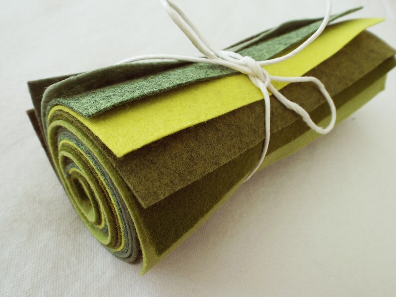 Puede incluir: Un rollo de tela de fieltro en tonos verdes y amarillos, atado con una cuerda blanca. El fieltro est&aacute; en capas en diferentes tonos de verde y amarillo, creando un efecto degradado.