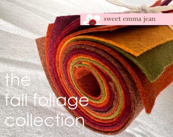 May include: A rolled bundle of felt sheets in autumn colors including deep red, orange, yellow, and olive green, tied with white string. A pink label features a flower icon and the text sweet emma jean, with the text the fall foliage collection.