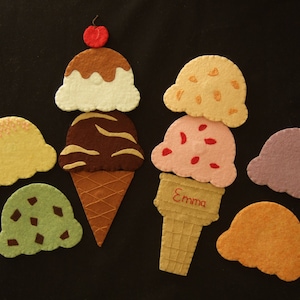Könnte beinhalten: Ein Set aus Filz-Eiswaffeln, darunter eine Schokoladen-, Erdbeer-, Vanille- und eine grüne Minzeiswaffel. Außerdem gibt es eine gelbe Zitroneneiswaffel, eine orangefarbene Sorbet-Eiswaffel und eine lila Trauben-Eiswaffel. Eine der Waffeln trägt den Namen "Emma" gestickt.
