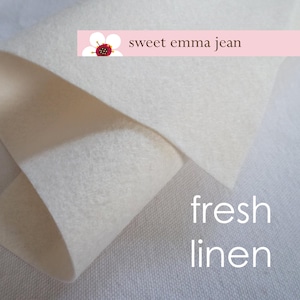May include: Close-up of two pieces of white fabric. The top piece is folded over the bottom piece. The top piece is a soft, white felt fabric. The bottom piece is a textured, white linen fabric. The text "sweet emma jean" and "fresh linen" is printed on the fabric.
