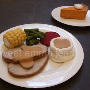 以下が含まれることがあります： 白いお皿にのったフェルト製の食べ物セット。カボチャパイ、トウモロコシ、インゲン、赤いビーツ、茶色のフィリングが入ったロールパン、ソースのかかった肉のスライス2枚が含まれています。「sweet emma jean」の文字が見えます。
