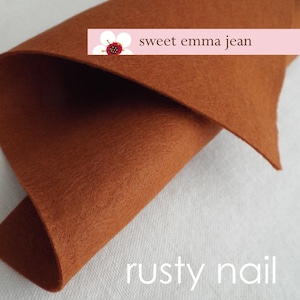 Puede incluir: Primer plano de un trozo de tela de fieltro de color naranja oxidado. La tela está doblada por la mitad y se puede ver el texto "sweet emma jean" y "rusty nail".