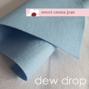 Könnte beinhalten: Ein hellblaues Filztuch, in der Hälfte gefaltet, mit dem Text "sweet emma jean" und "dew drop" auf der weißen Oberfläche darunter.