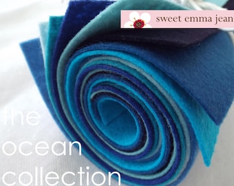 May include: A roll of felt fabric in shades of blue, teal, and white. The felt is rolled up tightly and the ends are visible. The text "sweet emma jean" is printed on a white tag attached to the roll. The text "the ocean collection" is printed below the roll of felt.