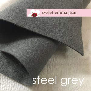 May include: A close-up of a folded piece of steel grey felt fabric. The fabric is soft and textured. The text "sweet emma jean" is printed on a pink label above the fabric.