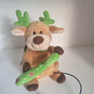 Può includere: Un giocattolo di peluche a forma di renna con pelliccia marrone, corna verdi e una sciarpa verde. La renna tiene in mano un oggetto verde cucito con dettagli rossi. Un cavo nero si estende dal giocattolo.