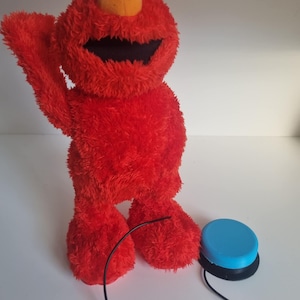 Puede incluir: Un peluche rojo de Elmo con ojos blancos grandes, una nariz naranja y una boca muy abierta. Tiene un botón azul conectado por un cable negro. El juguete está sobre una superficie blanca.