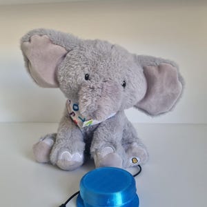 Puede incluir: Un peluche de elefante gris con orejas grandes y una bufanda estampada. Un botón azul circular con un cable negro está delante del elefante. El juguete tiene un botón en su pie.