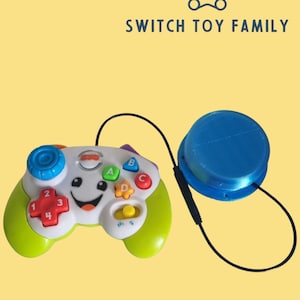 May include: A white and green toy game controller with a smiling face, colorful buttons labeled 1, 2, 3, 4, A, B, C, D, and a blue circular button connected by a black wire. The text SWITCH TOY FAMILY appears above with a bear icon.