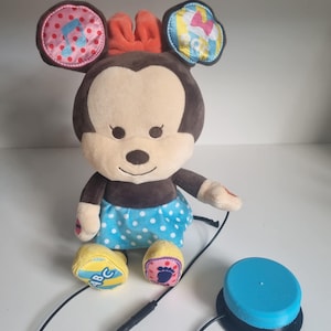 May include: A plush Minnie Mouse toy with a blue polka dot skirt, a red bow, and patterned ears. The toy is connected to a blue button with a black cord. The ears have musical notes, a bow, and the ABCs.