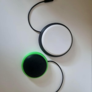 May include: Two circular push-button switches with black cables and 3.5mm audio jacks. One switch features a white top with a black base, while the other switch features a black top with a bright green base. Both devices are round and flat.