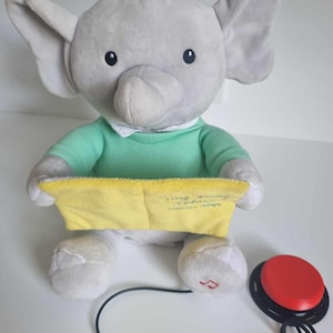 May include: A grey plush elephant toy wearing a green shirt and holding a yellow book. The elephant has a red button on its foot and is connected to a red button on a black cord. The book says "Sleepy Time Stories" on the front.