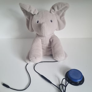 May include: A plush gray elephant toy with large ears and small black eyes. The elephant is connected to a black wire and a blue button. The toy is designed for interactive play and sensory engagement.