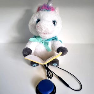 Puede incluir: Un juguete de peluche de unicornio blanco con un cuerno plateado y una melena colorida. El unicornio lleva una bufanda azul y sostiene un pequeño libro. Un botón azul está conectado al juguete por un cable negro.