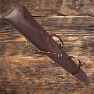 May include: A brown leather gun case with a shoulder strap and a handle. The case has a zipper closure and a small logo on the front.