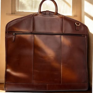 Puede incluir: Una funda de ropa de cuero marrón con un asa superior y una cremallera de cuerpo entero. La bolsa tiene forma rectangular con detalles de costura vertical y un pequeño anillo de metal en el lateral. Diseñada para llevar trajes y otras prendas.