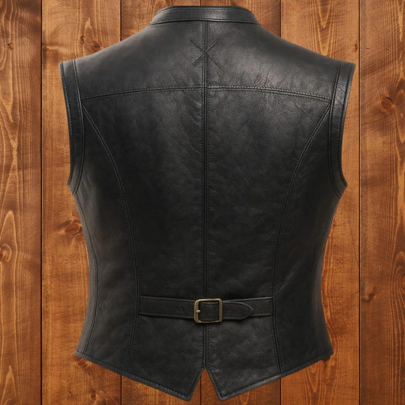 May include: A black leather vest with a tailored fit, featuring a collarless design and a buckle closure at the waist. The vest has a classic, timeless style, with visible stitching details and a smooth, matte finish. The background is a wooden surface.