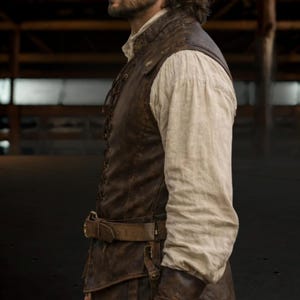 Op de afbeelding: Een bruin leren vest met een veter aan de voorkant en een bijpassende riem. Het vest wordt gedragen over een wit shirt met lange mouwen. De outfit heeft een rustieke, historische uitstraling.