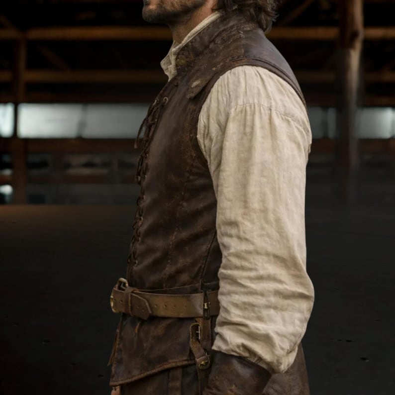 Op de afbeelding: Een bruin leren vest met een veter aan de voorkant en een bijpassende riem. Het vest wordt gedragen over een wit shirt met lange mouwen. De outfit heeft een rustieke, historische uitstraling.