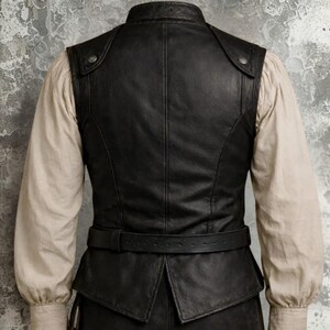 Op de afbeelding: Zwart leren vest met een hoge kraag en een riem. Het vest heeft een getailleerde pasvorm met knoopdetails op de schouders. Het vest wordt gedragen over een cr&egrave;mekleurig shirt met lange mouwen.