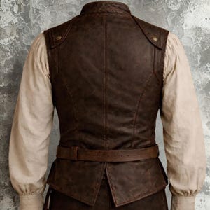 Op de afbeelding: Een bruin leren vest met een hoge kraag en een riem. Het vest heeft een verweerde uitstraling en wordt gedragen over een cr&egrave;mekleurig shirt met lange mouwen. De achterkant van het vest is zichtbaar, met stikseldetails en een getailleerde pasvorm.