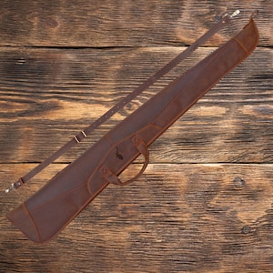 May include: A brown leather gun case with a shoulder strap and a handle. The case has a decorative design on the front.