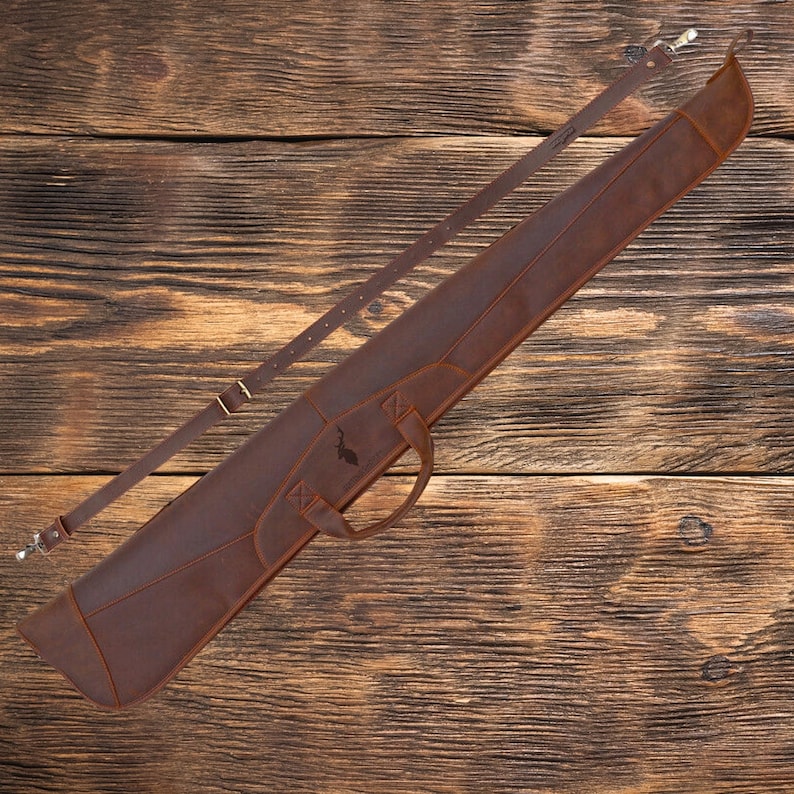 May include: A brown leather gun case with a shoulder strap and a handle. The case has a decorative design on the front.